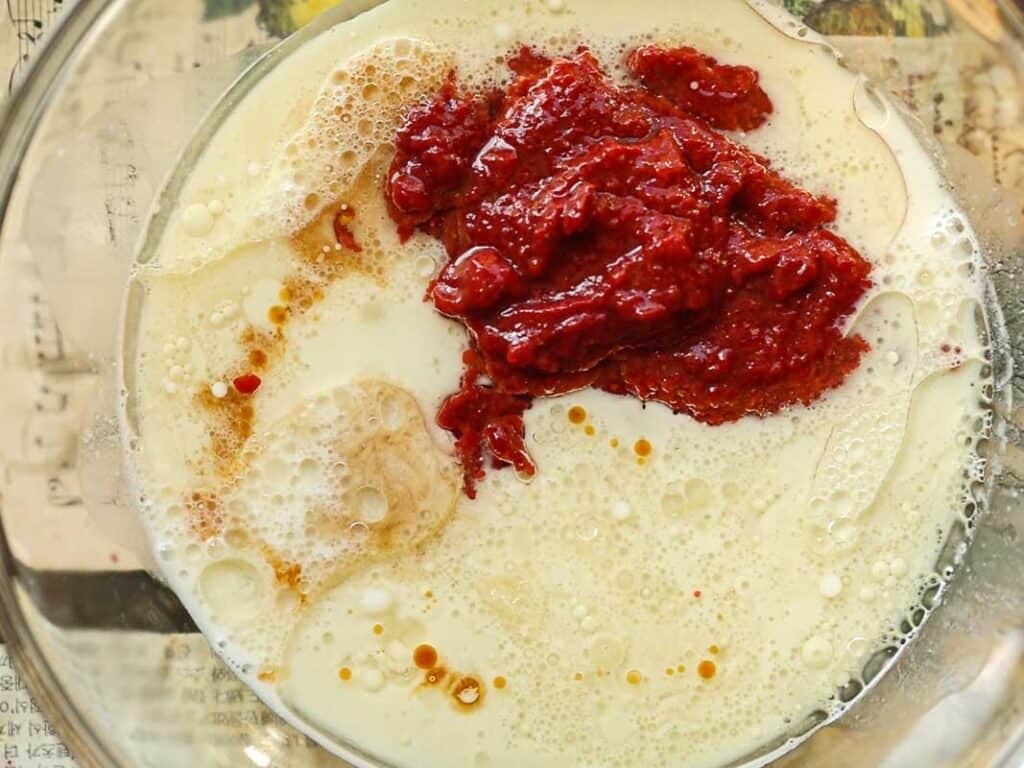 A glass bowl containing milk, strawberry reduction, and other ingredients for strawberry cake batter. The ingredients are partially mixed, with some floating bubbles visible. The bowl is placed on a beige surface with faint text.